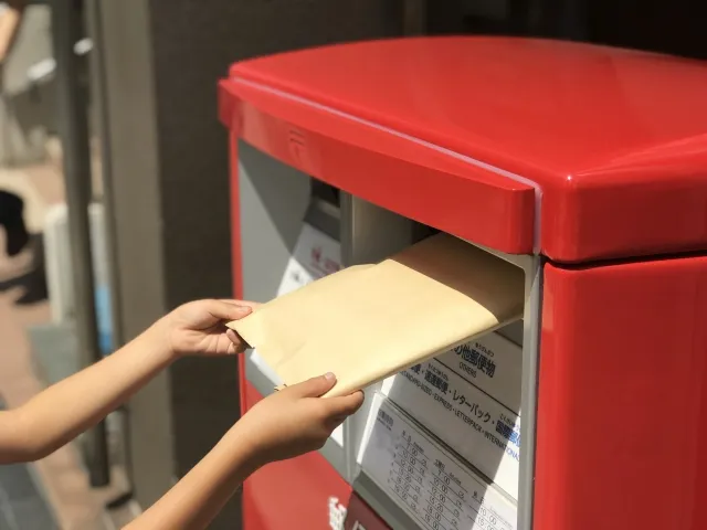 クリックポストのラベルを貼った茶封筒を、街中の赤い郵便ポストの投入口へ差し入れている手元のアップ写真。クリックポストがいつでもポストから手軽に発送できることを表現しています。