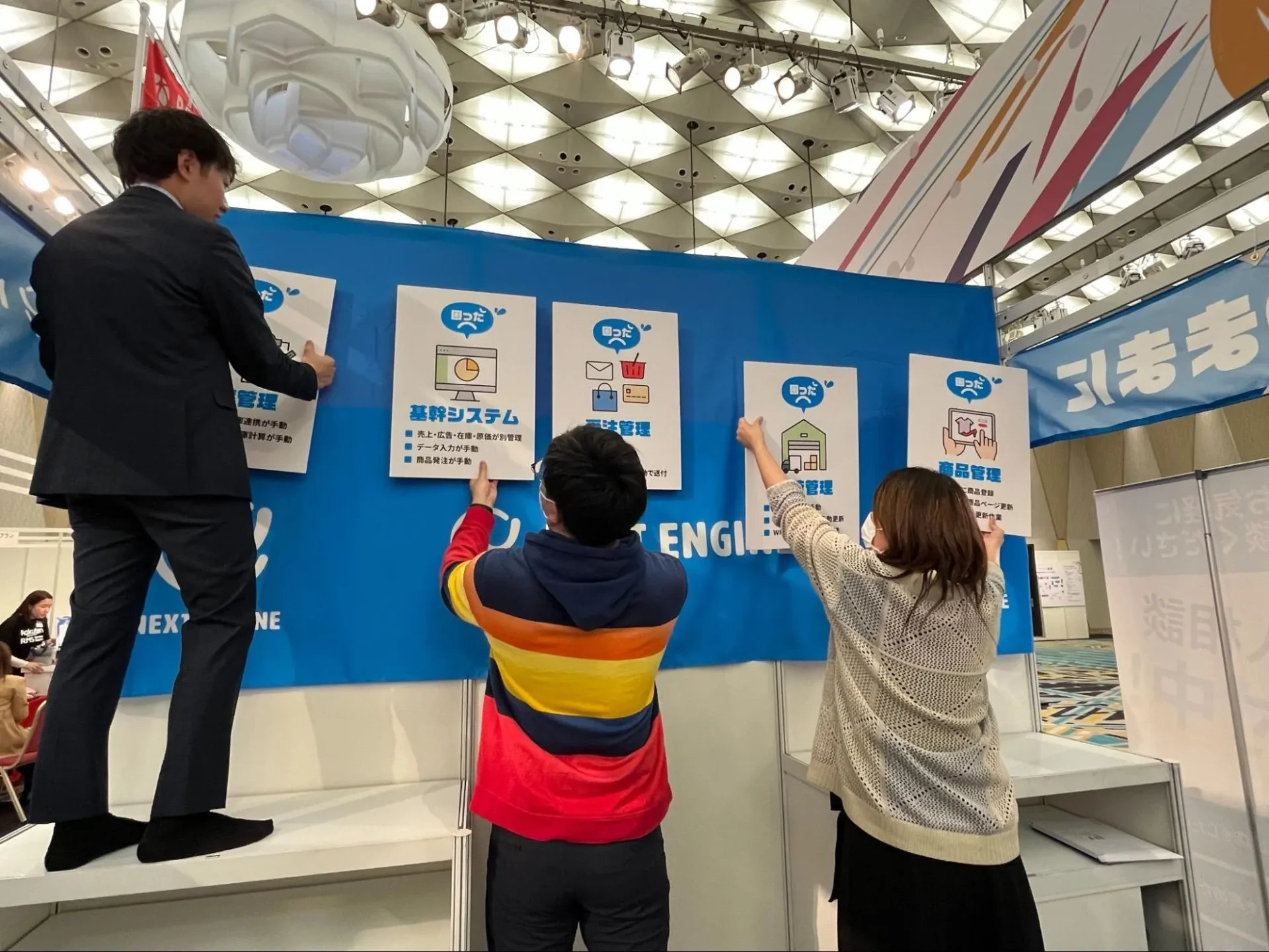 ネクストエンジンブースの壁面に、基幹システムや受注管理、商品管理などの「お悩み解決パネル」を設置する設営スタッフの様子。