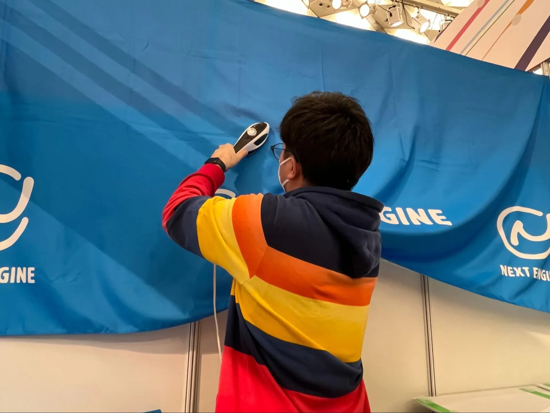 ネクストエンジンブースの設営風景。青いブランドバナーのシワをスチーマーで丁寧に伸ばしているスタッフの背中。