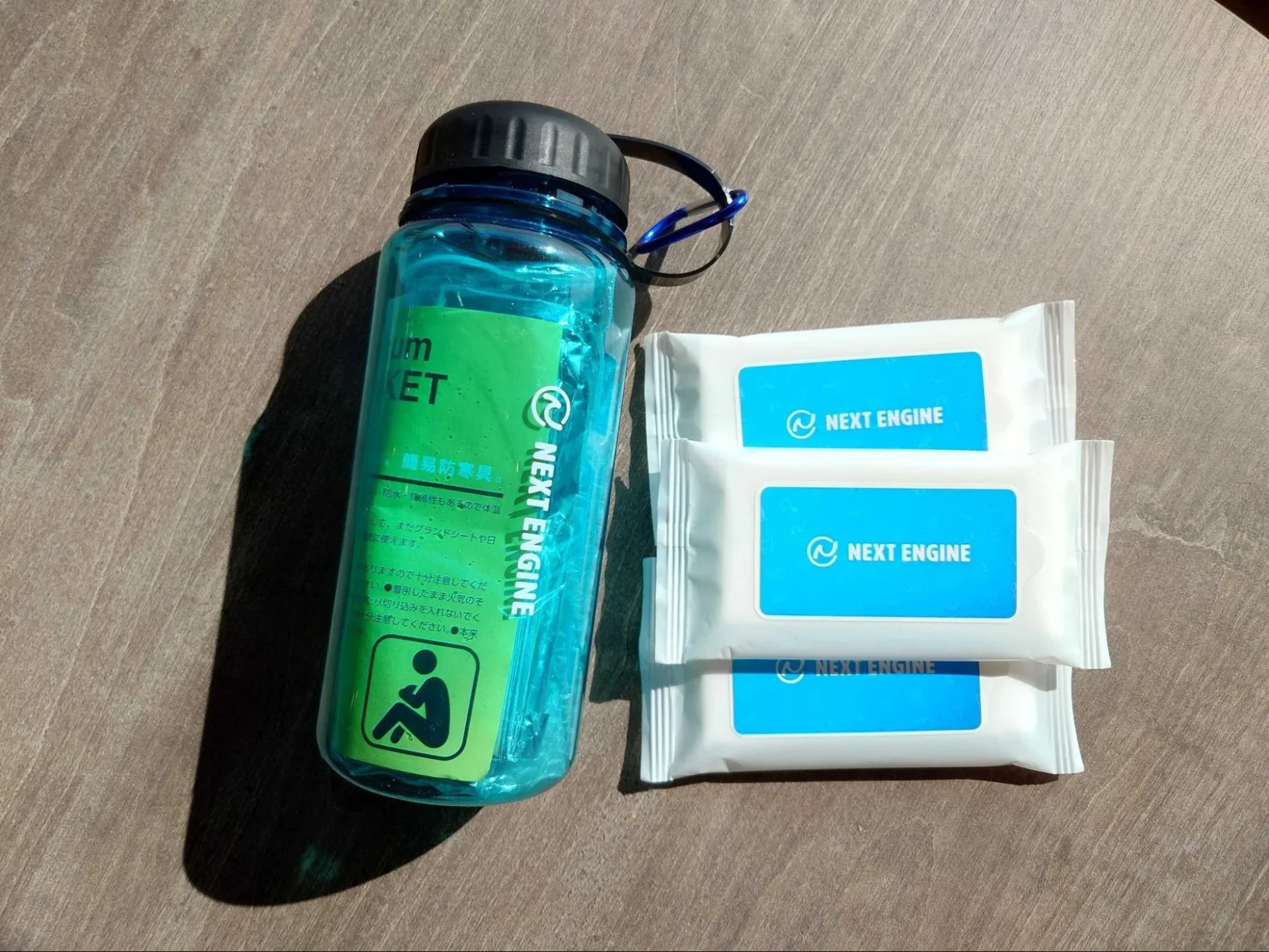 ネクストエンジンのロゴが入った、ノベルティの青いクリアボトルとウェットティッシュの俯瞰写真。
